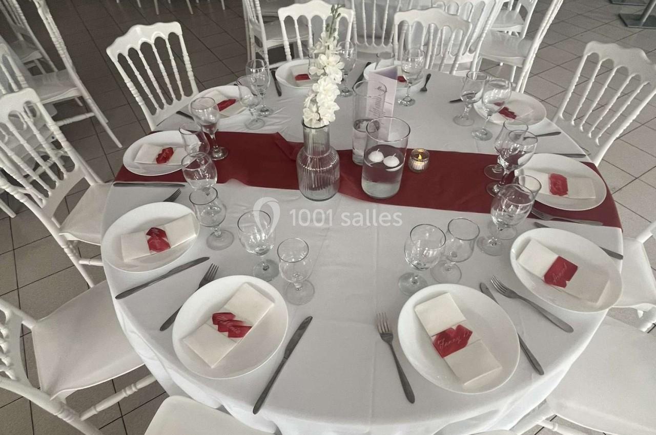 Table ronde dressée avec nappes blanches, chemin de table rouge, vaisselle, verres et pliages de serviettes rouges.