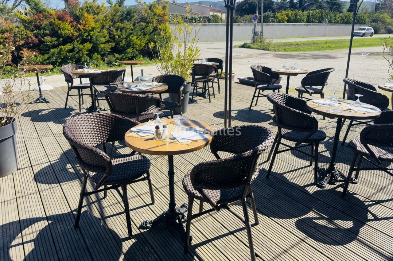 Terrasse ensoleillée avec tables rondes en bois, chaises en osier et couverts disposés, entourée de verdure.