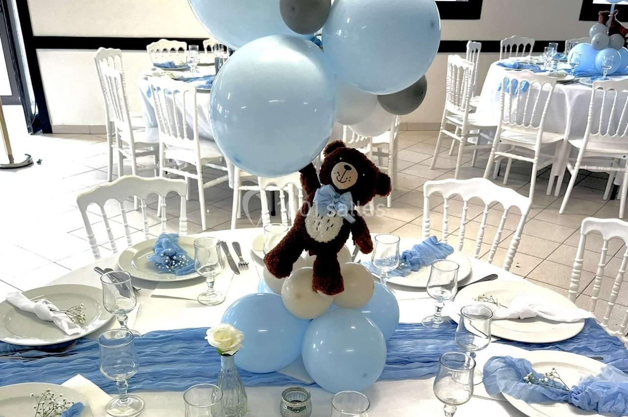 Centre de table avec ballons bleus et blancs, ourson en peluche, nappes bleues et vaisselle disposée sur une table.
