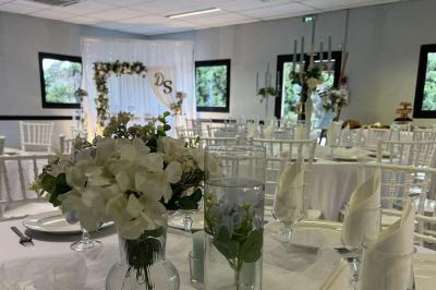 Salle de réception décorée avec des tables rondes dressées, nappes blanches, chaises blanches et touches de rose.