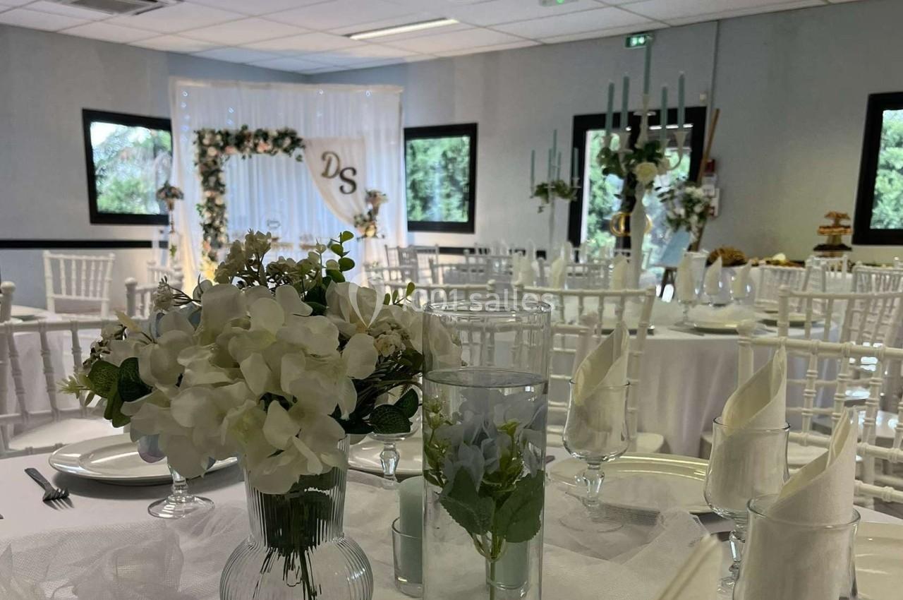 Salle de réception décorée pour un mariage avec des tables dressées, des fleurs blanches et une arche florale en arrière…