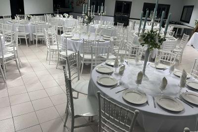 Salle de réception décorée avec des tables rondes dressées, nappes blanches, chaises blanches et touches de rose.