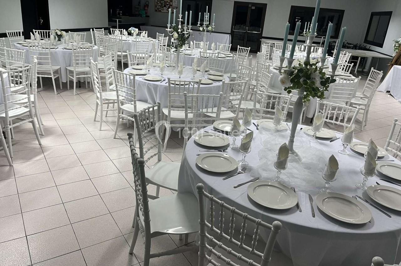 Salle de réception avec des tables rondes dressées, nappes blanches, chaises blanches et centres de table décoratifs.