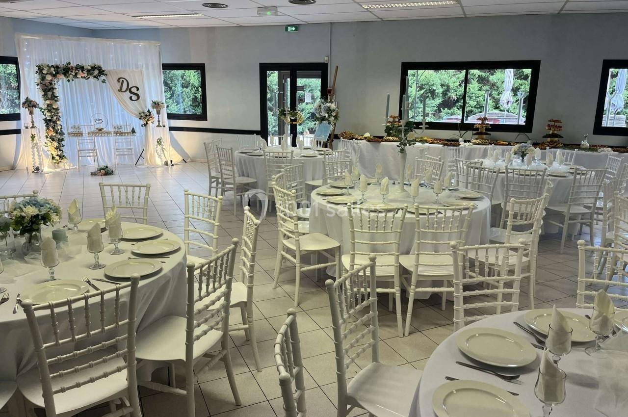 Salle de réception décorée pour un mariage, avec des tables rondes dressées et une arche florale en arrière-plan.