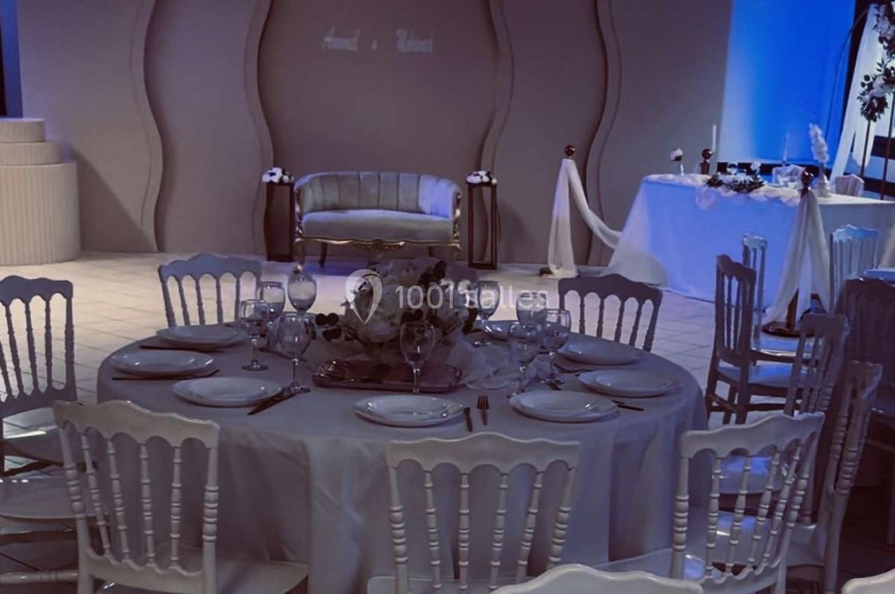 Salle de réception décorée avec des tables rondes dressées, des chaises blanches et un espace central avec canapé.
