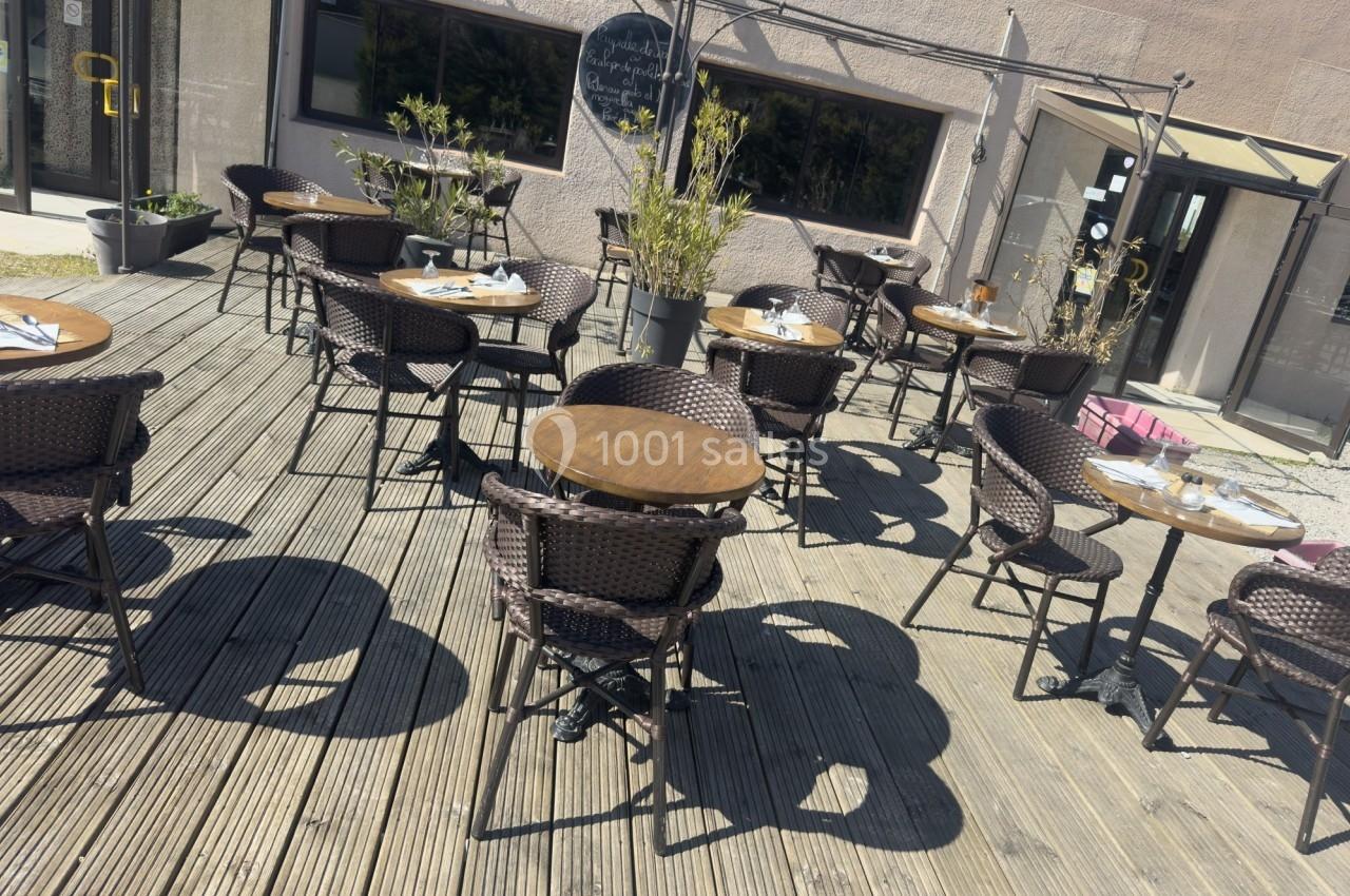 Terrasse en bois avec des tables rondes et des chaises en osier, partiellement ombragée par des plantes et des parasols.