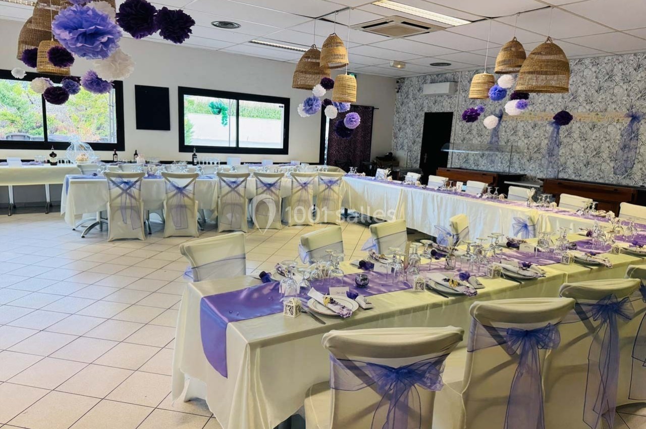 Salle de réception décorée avec des tables dressées, nappes blanches et accents violets, prête pour un événement.