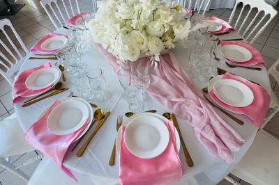 Salle de réception décorée avec des tables rondes dressées, nappes blanches, chaises blanches et touches de rose.