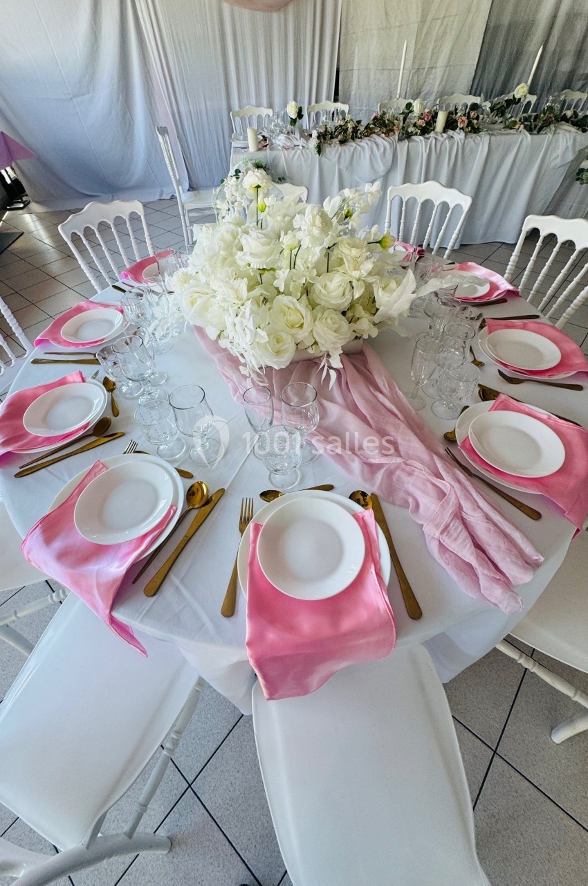 Table ronde décorée avec une nappe blanche, serviettes roses, couverts dorés et un centre de table floral blanc.