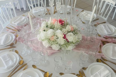 Salle de réception décorée avec des tables rondes dressées, nappes blanches, chaises blanches et touches de rose.