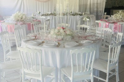 Salle de réception décorée avec des tables rondes dressées, nappes blanches, chaises blanches et touches de rose.
