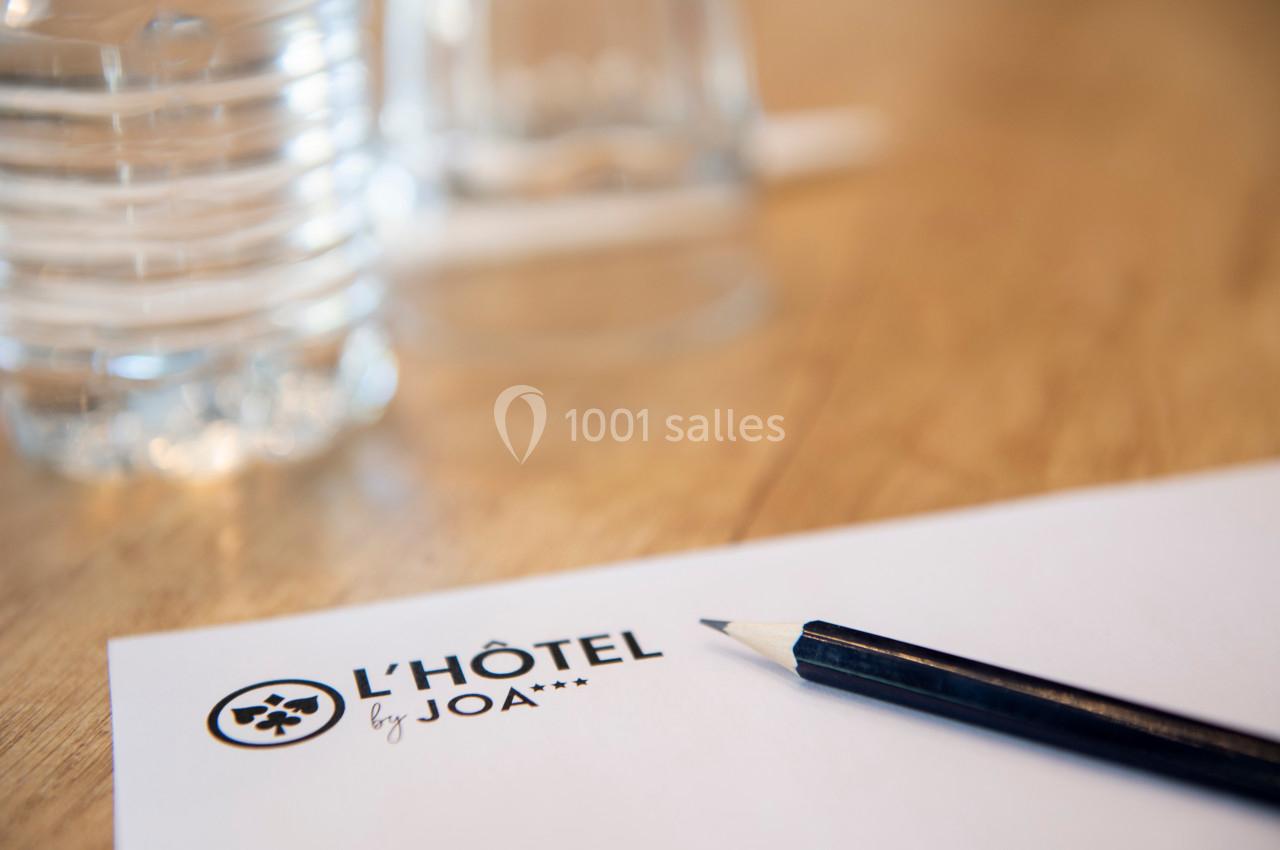 Feuille avec le logo ’L'Hôtel by JOA’, un crayon noir et une bouteille d'eau sur une table en bois.