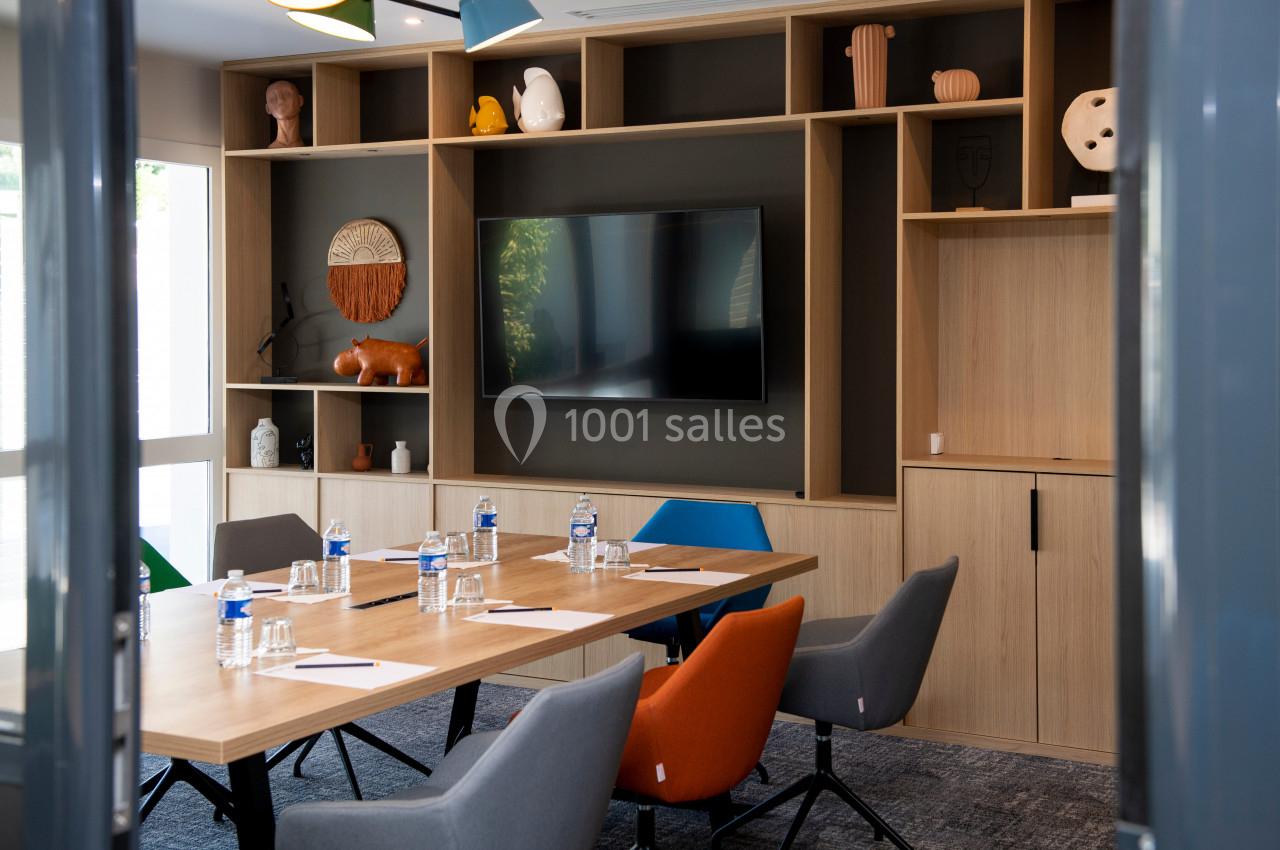 Salle de réunion moderne avec table en bois, chaises colorées, écran mural et étagères décoratives en arrière-plan.