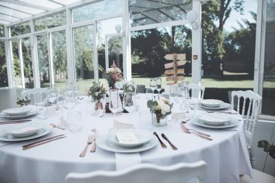 Salle de réception lumineuse avec tables dressées, lustres suspendus et nappes blanches sur des tables latérales.