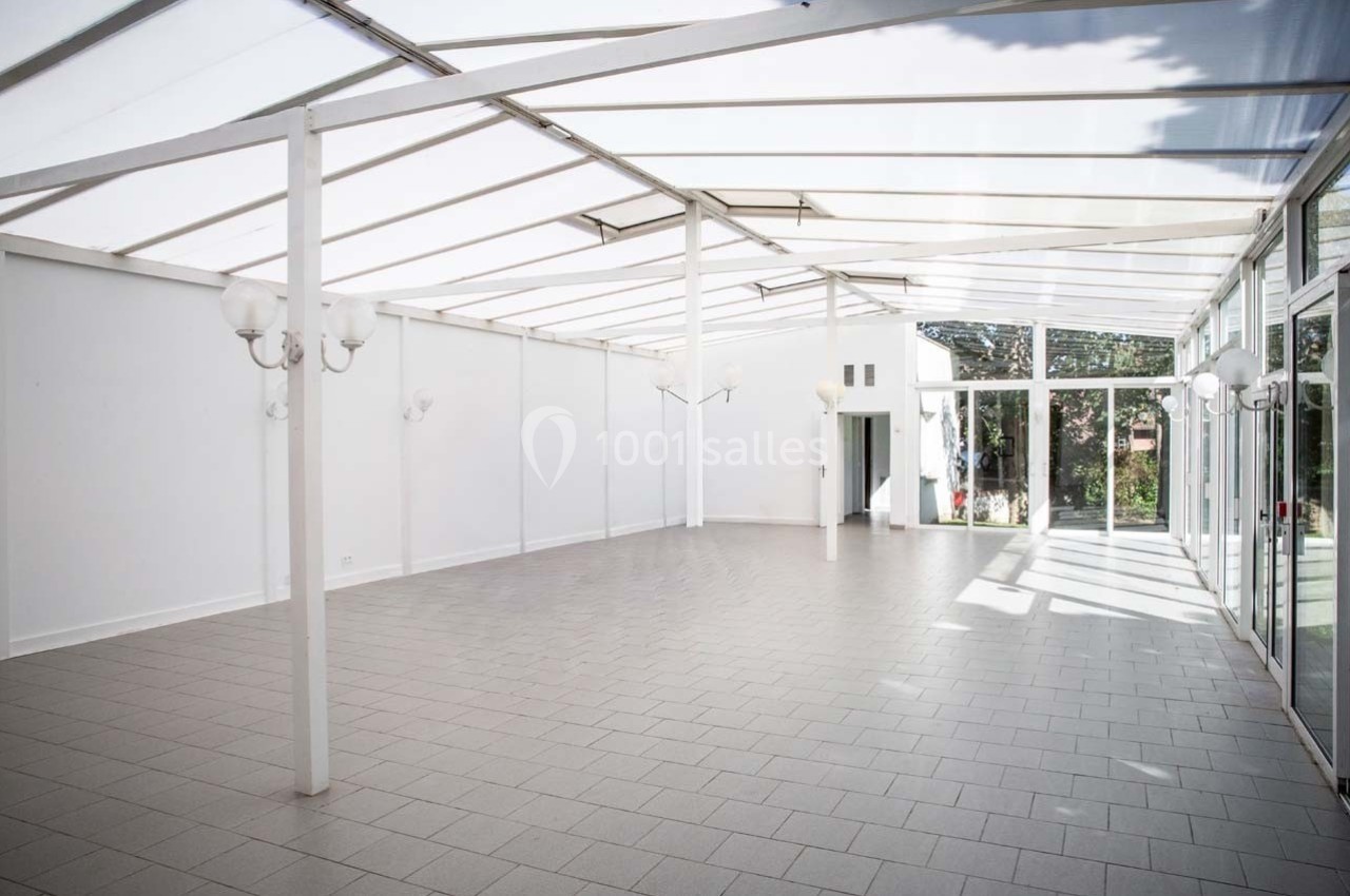 Salle lumineuse avec toit en verre, murs blancs, sol carrelé gris et grandes baies vitrées donnant sur un jardin.