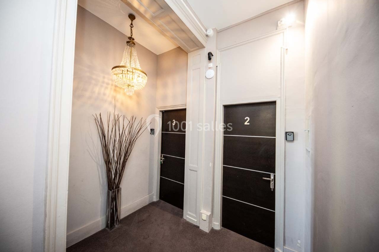 Couloir intérieur avec deux portes numérotées 2 et 3, un lustre suspendu et un vase décoratif rempli de branches.