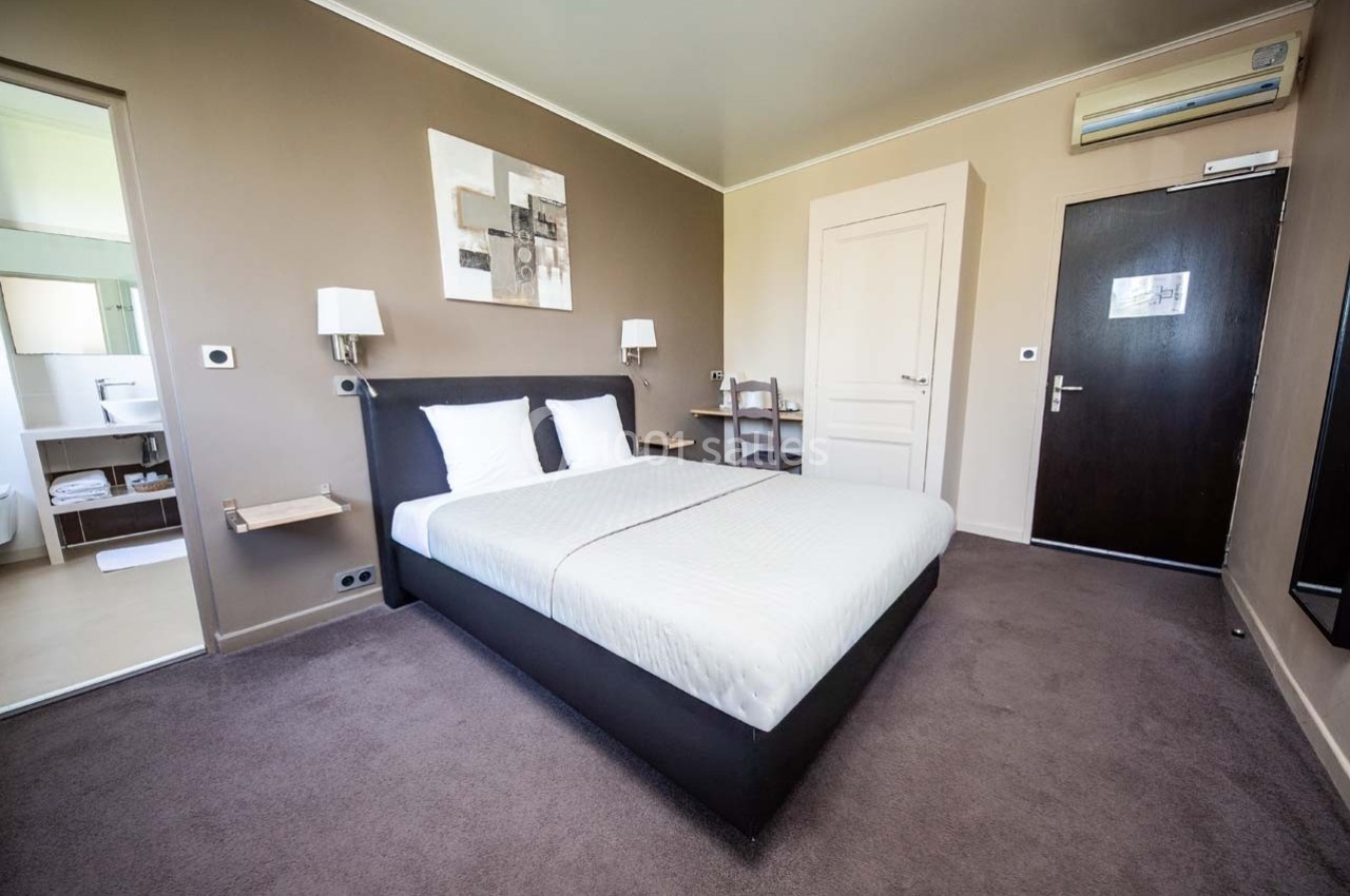 Chambre d'hôtel avec lit double, murs beige, bureau, chaise, porte noire et salle de bain visible en arrière-plan.