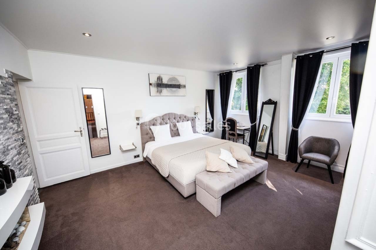 Chambre lumineuse avec lit double, fauteuil, miroirs, rideaux noirs et fenêtres donnant sur un espace vert.