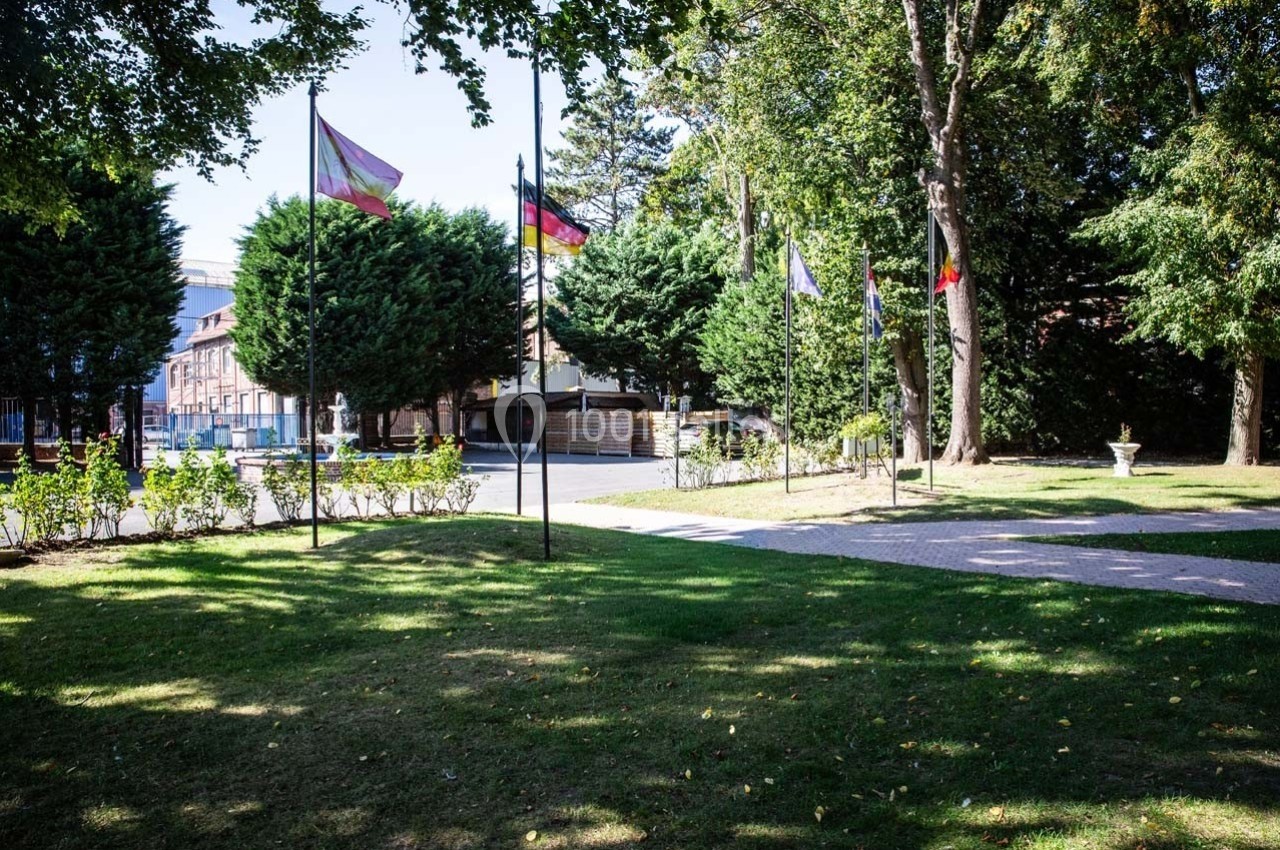 Pelouse avec arbres, allée pavée et drapeaux sur mâts devant des bâtiments en arrière-plan.