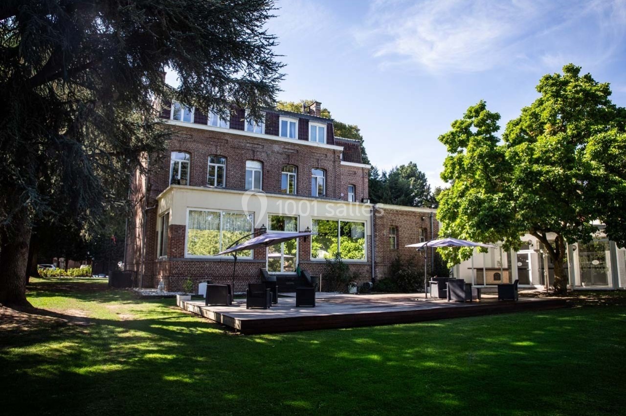 Façade d'un bâtiment en briques entouré d'arbres, avec une terrasse en bois aménagée de tables et parasols.