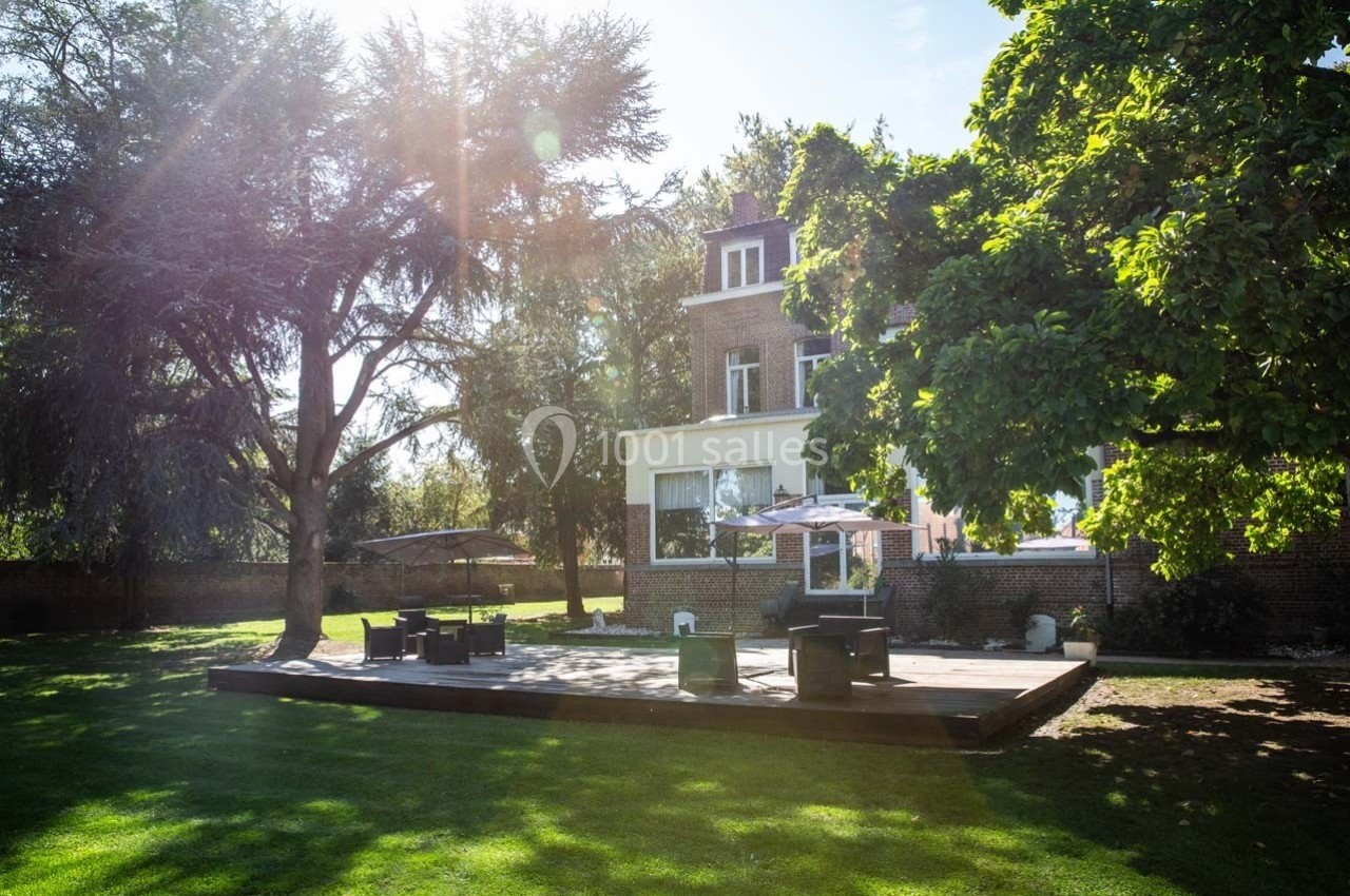 Terrasse en bois avec mobilier de jardin, entourée d'arbres et située devant une maison en briques sous un ciel ensoleillé.