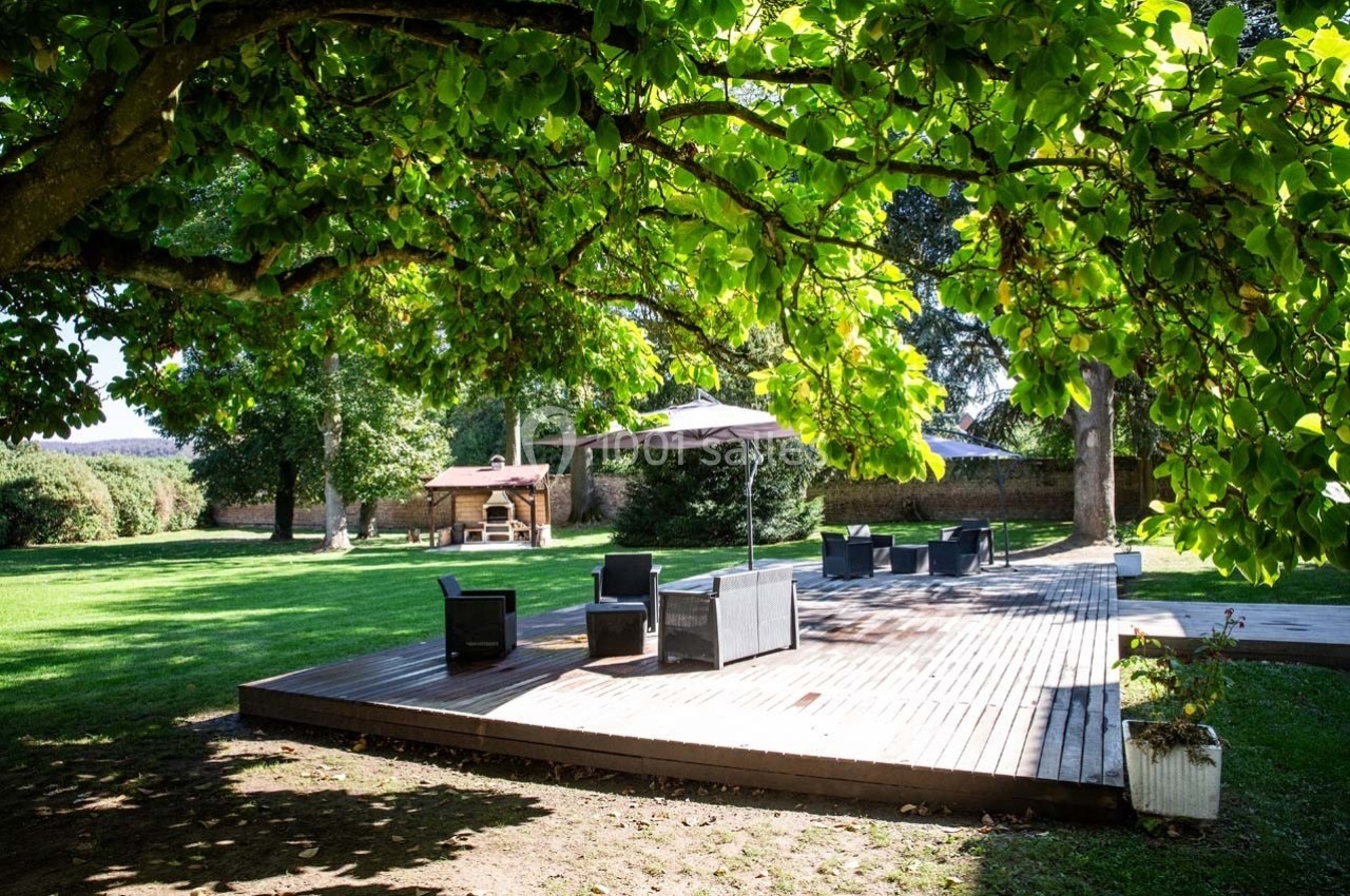 Terrasse en bois avec mobilier de jardin sous un grand arbre, entourée de pelouse et d'arbres dans un espace extérieur.