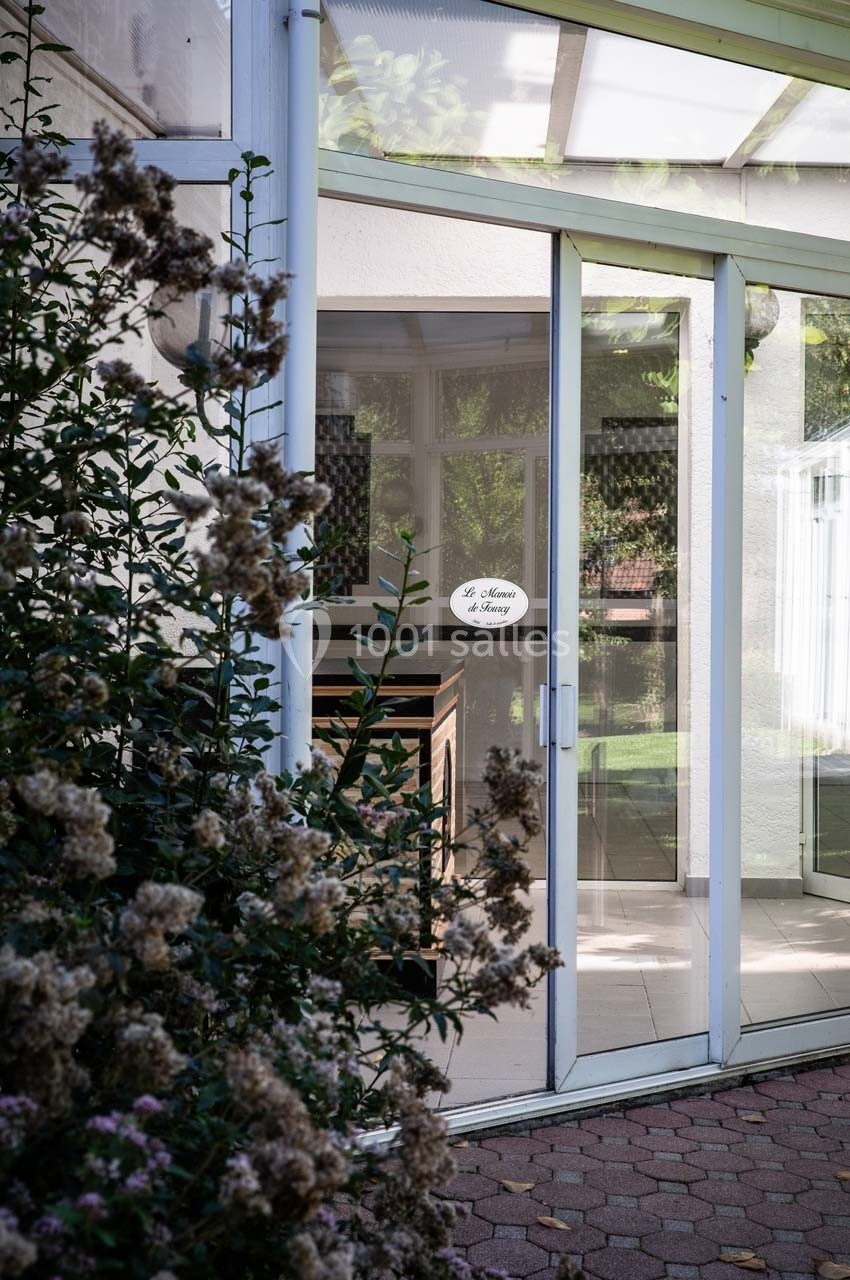 Vue d'une porte vitrée donnant sur un intérieur lumineux, entourée de végétation et d'un chemin en pavés.