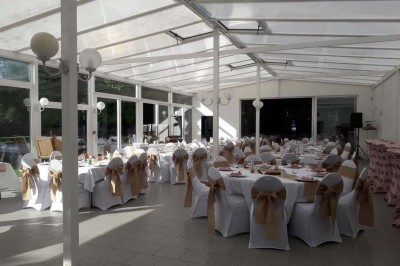 Salle de réception lumineuse avec tables dressées, lustres suspendus et nappes blanches sur des tables latérales.