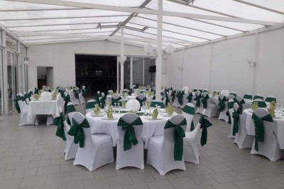 Salle de réception lumineuse avec tables dressées, lustres suspendus et nappes blanches sur des tables latérales.