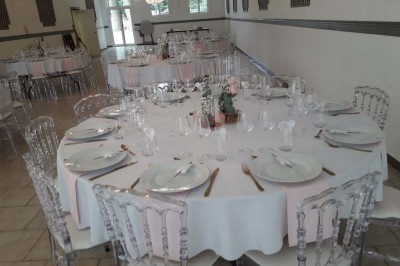 Salle de réception lumineuse avec tables dressées, lustres suspendus et nappes blanches sur des tables latérales.