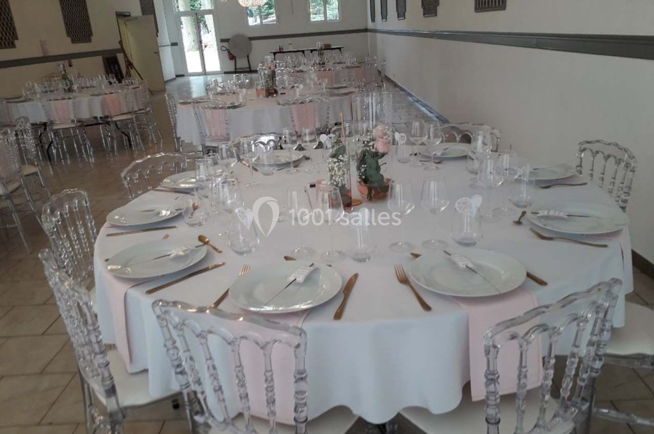 Salle de réception avec tables rondes dressées, nappes blanches, chaises transparentes et décoration florale centrale.