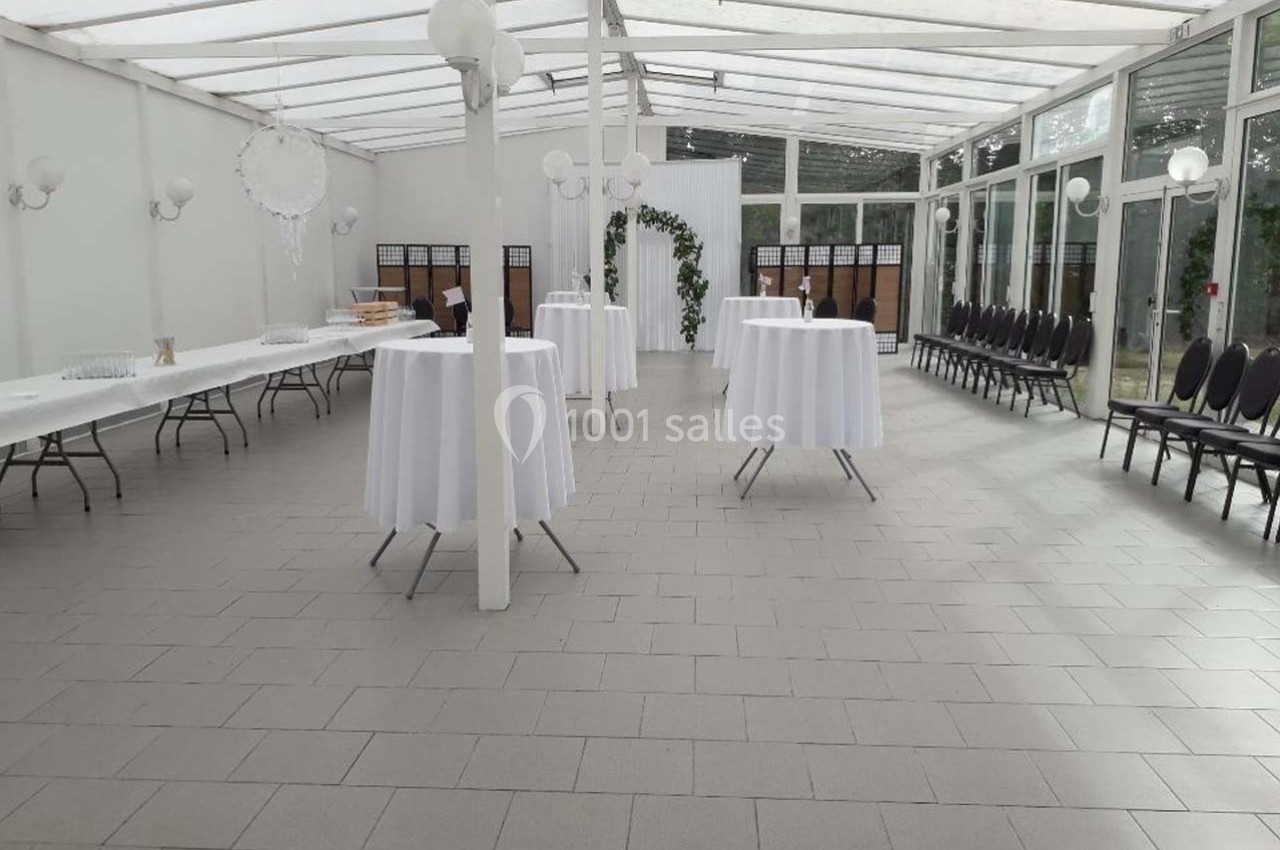 Salle lumineuse avec tables hautes drapées de blanc, chaises alignées et décoration végétale au fond.