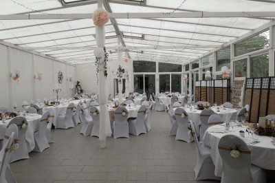 Salle de réception lumineuse avec tables dressées, lustres suspendus et nappes blanches sur des tables latérales.