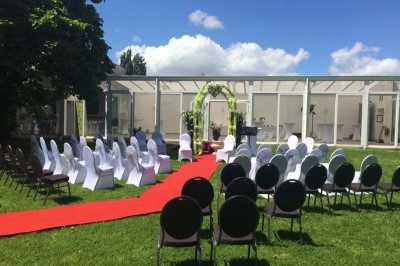 Salle de réception lumineuse avec tables dressées, lustres suspendus et nappes blanches sur des tables latérales.