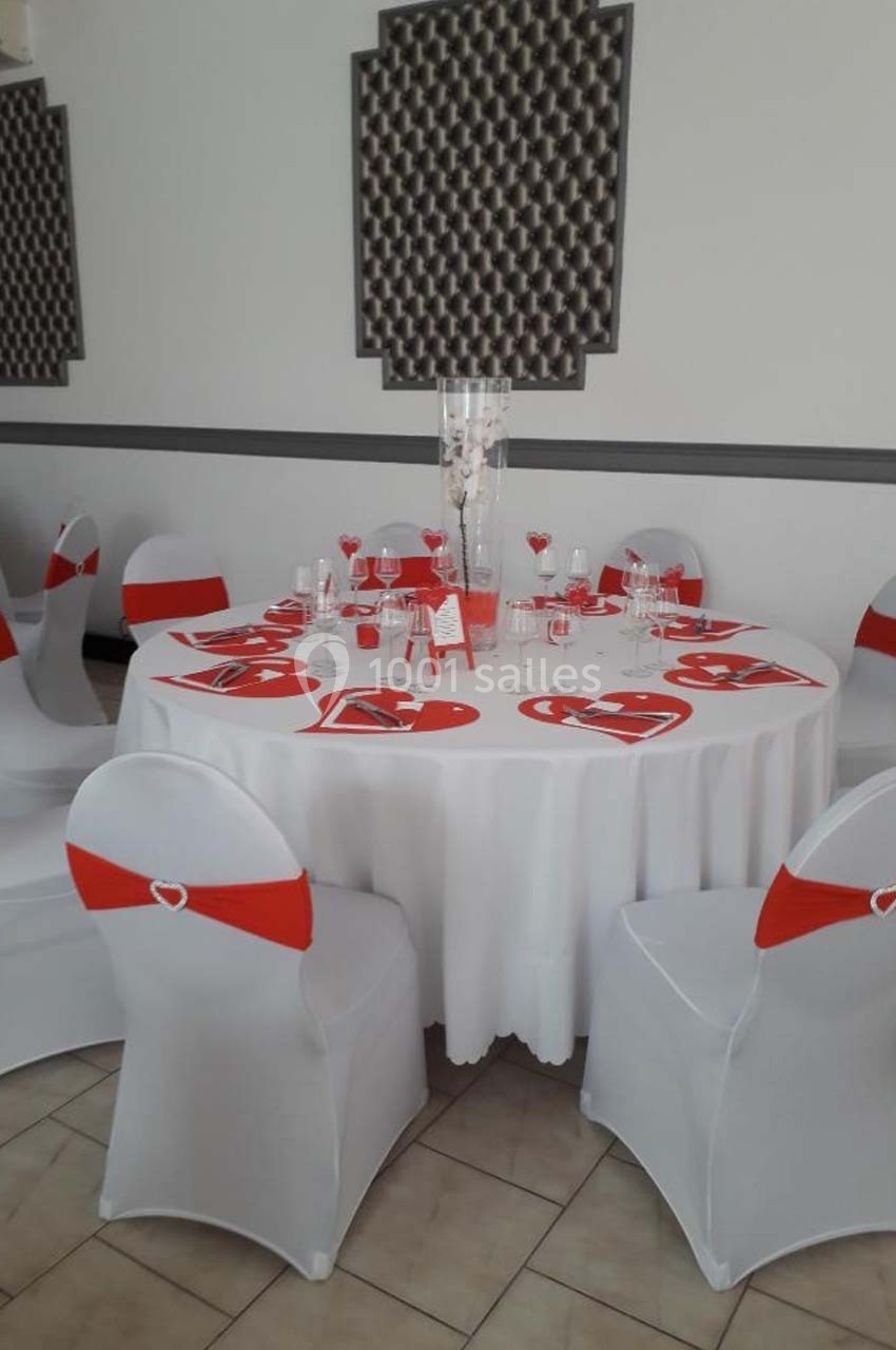 Table ronde décorée pour un événement, avec nappes blanches, chaises ornées de rubans rouges et vaisselle disposée.