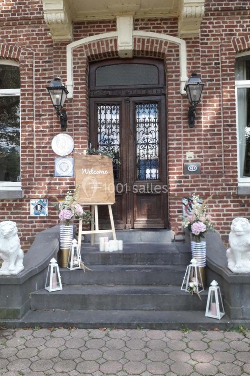 Entrée d'une maison en briques avec porte en bois, décorée de fleurs, lanternes et un panneau ’Welcome’.