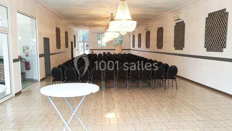 Salle de réunion avec rangées de chaises noires alignées, grandes fenêtres et lustres suspendus.