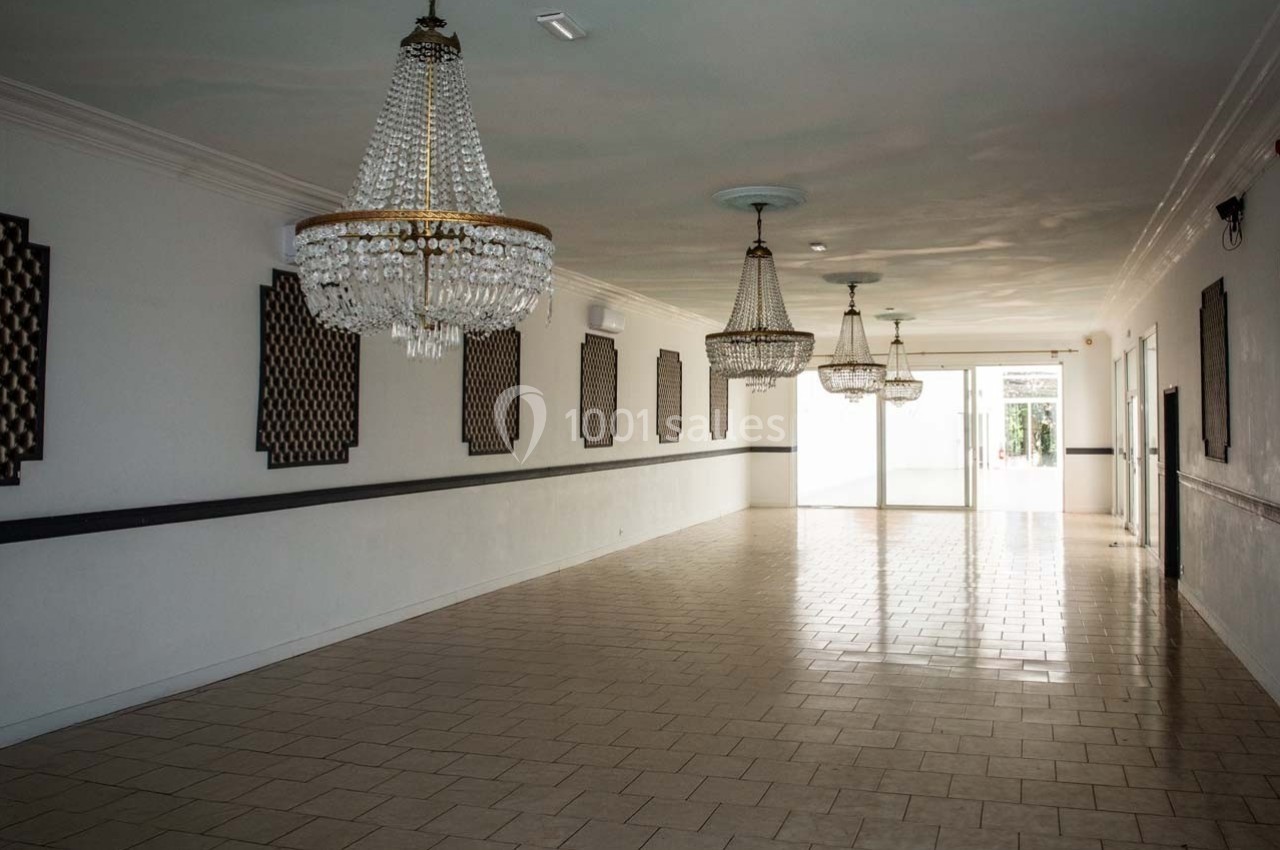 Salle vide avec carrelage clair, murs blancs, lustres en cristal suspendus et lumière naturelle entrant par une porte vitrée.