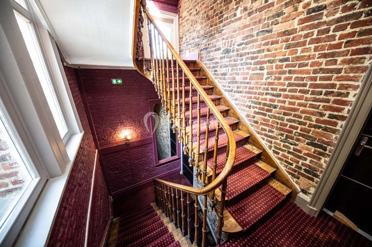 Escalier en bois avec rampe, tapis rouge et murs en briques, éclairé par une fenêtre et une applique murale.