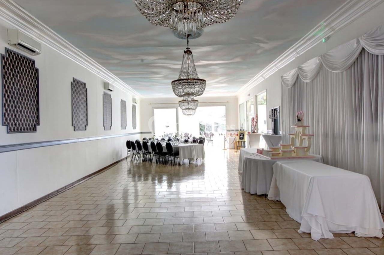 Salle de réception lumineuse avec tables dressées, lustres suspendus et nappes blanches sur des tables latérales.