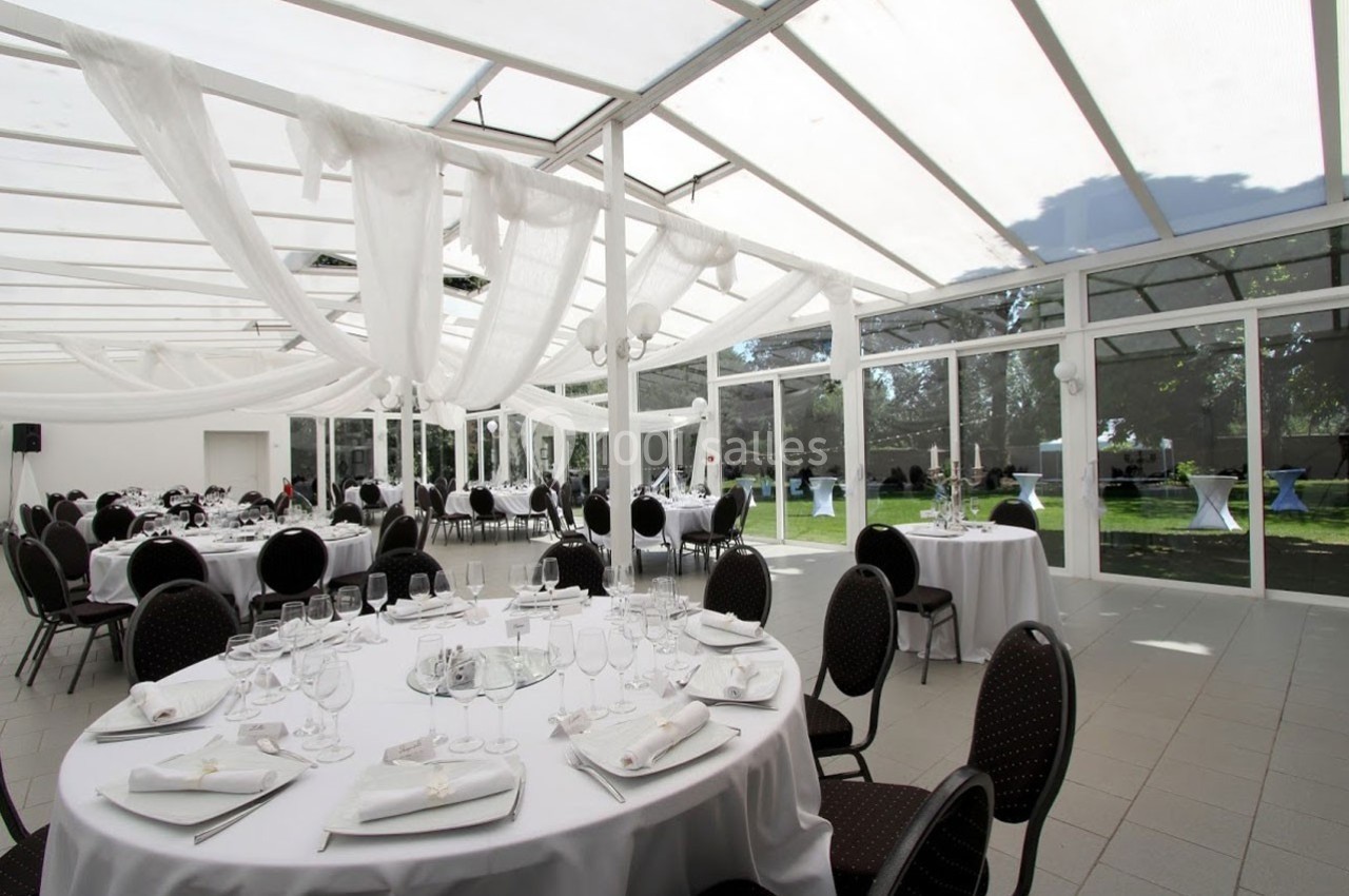 Salle de réception lumineuse avec tables rondes dressées, chaises noires et grandes baies vitrées donnant sur un jardin.
