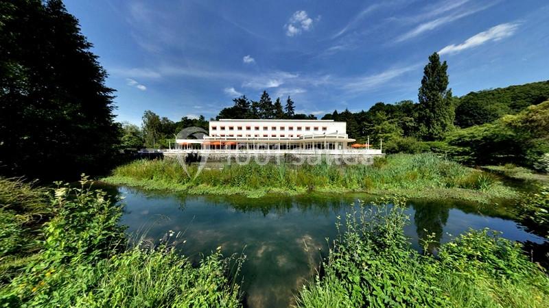 Bâtiment blanc moderne entouré de végétation, avec un plan d'eau au premier plan et un ciel bleu dégagé.