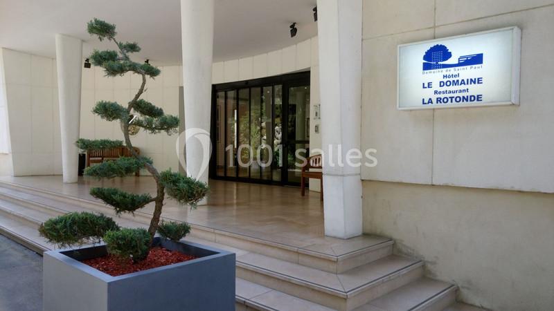 Entrée d'un hôtel avec une plante taillée en pot, des marches et une enseigne indiquant ’Le Domaine - La Rotonde’.