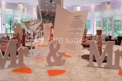 Salle lumineuse avec une grande table dressée pour un repas, décorée de fleurs et entourée de baies vitrées donnant sur un…