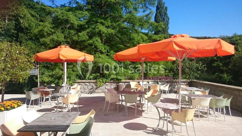 Terrasse ensoleillée avec tables, chaises et parasols orange, entourée de verdure et d'un ciel bleu dégagé.