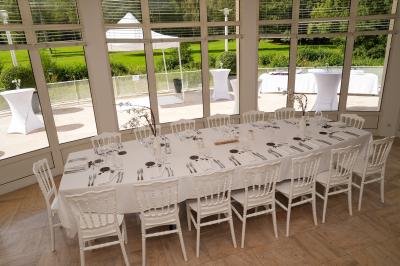 Salle lumineuse avec une grande table dressée pour un repas, décorée de fleurs et entourée de baies vitrées donnant sur un…