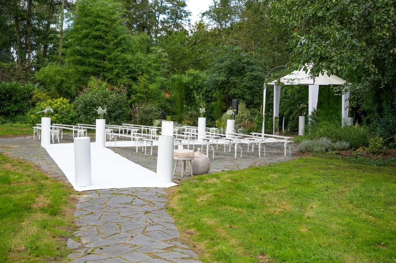 Allée en pierre menant à une cérémonie en extérieur avec bancs blancs, arche décorée et verdure environnante.