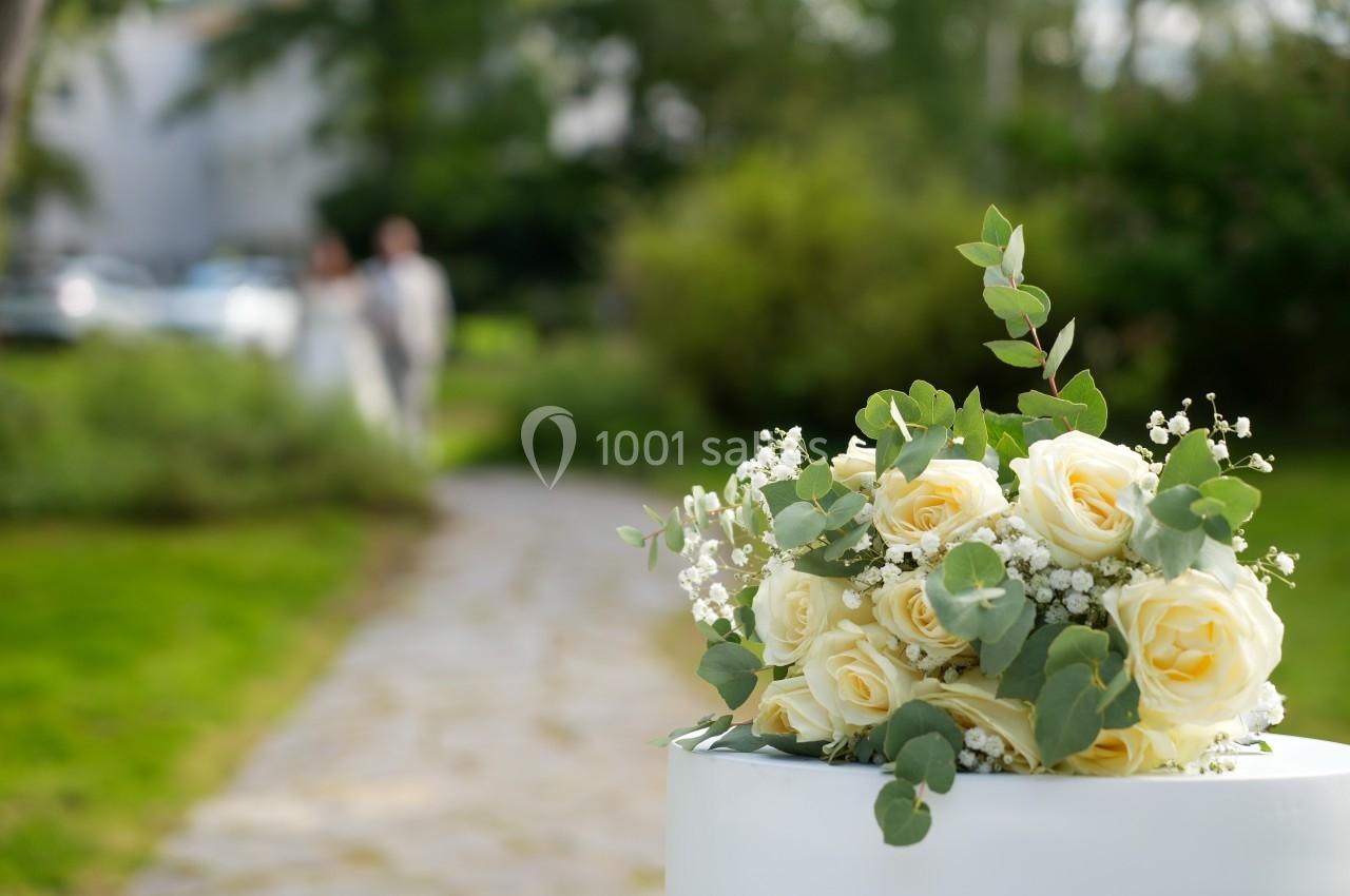 Bouquet de roses jaunes et de gypsophiles posé sur un support blanc, avec un couple flou en arrière-plan dans un jardin.