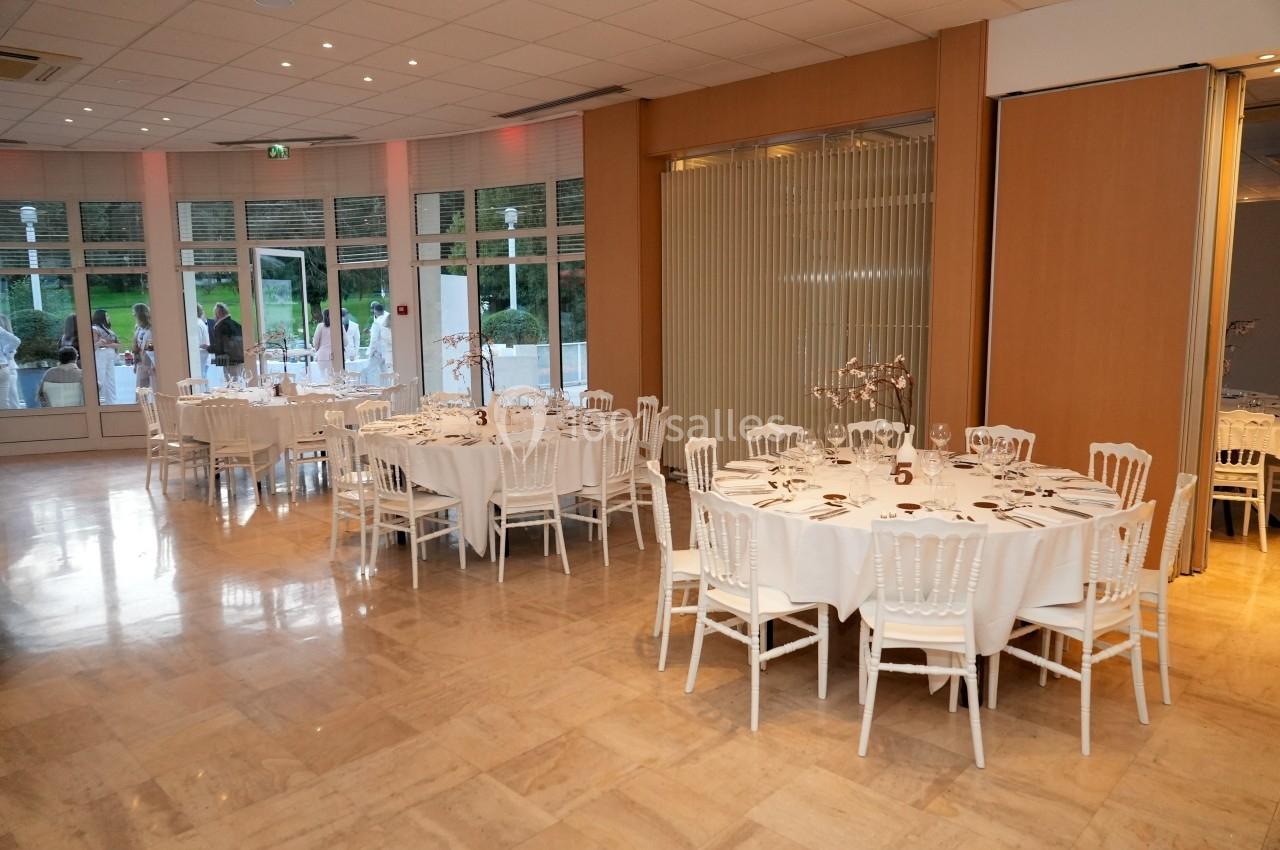 Salle de réception lumineuse avec tables rondes dressées, chaises blanches et vue sur un jardin à travers de grandes baies…