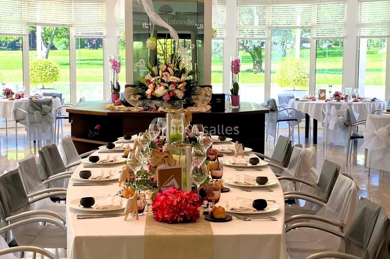 Salle lumineuse avec une grande table dressée pour un repas, décorée de fleurs et entourée de baies vitrées donnant sur un…